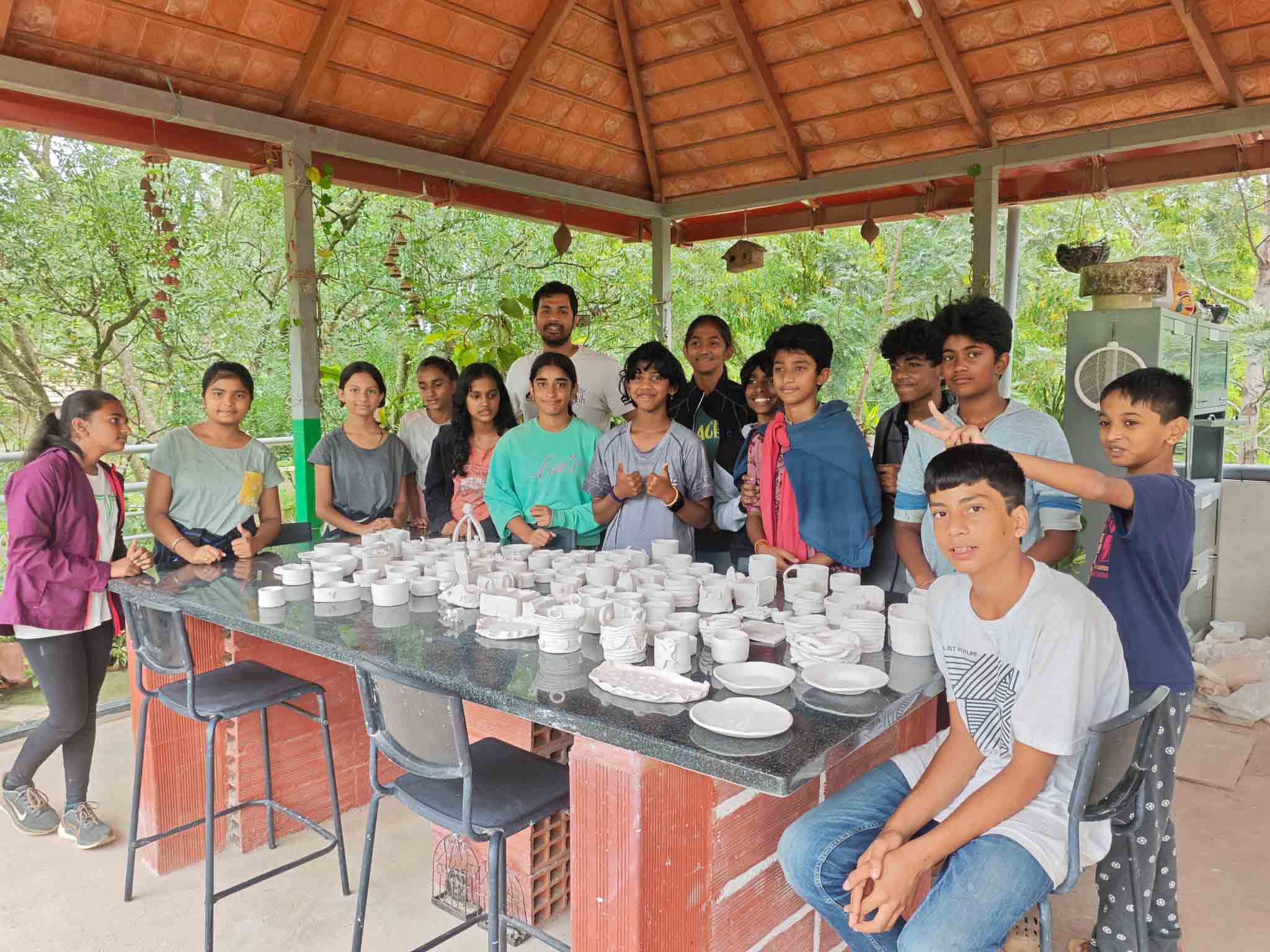 Young students around a table full of hand crafted pottery items in an outdoor studio setting.